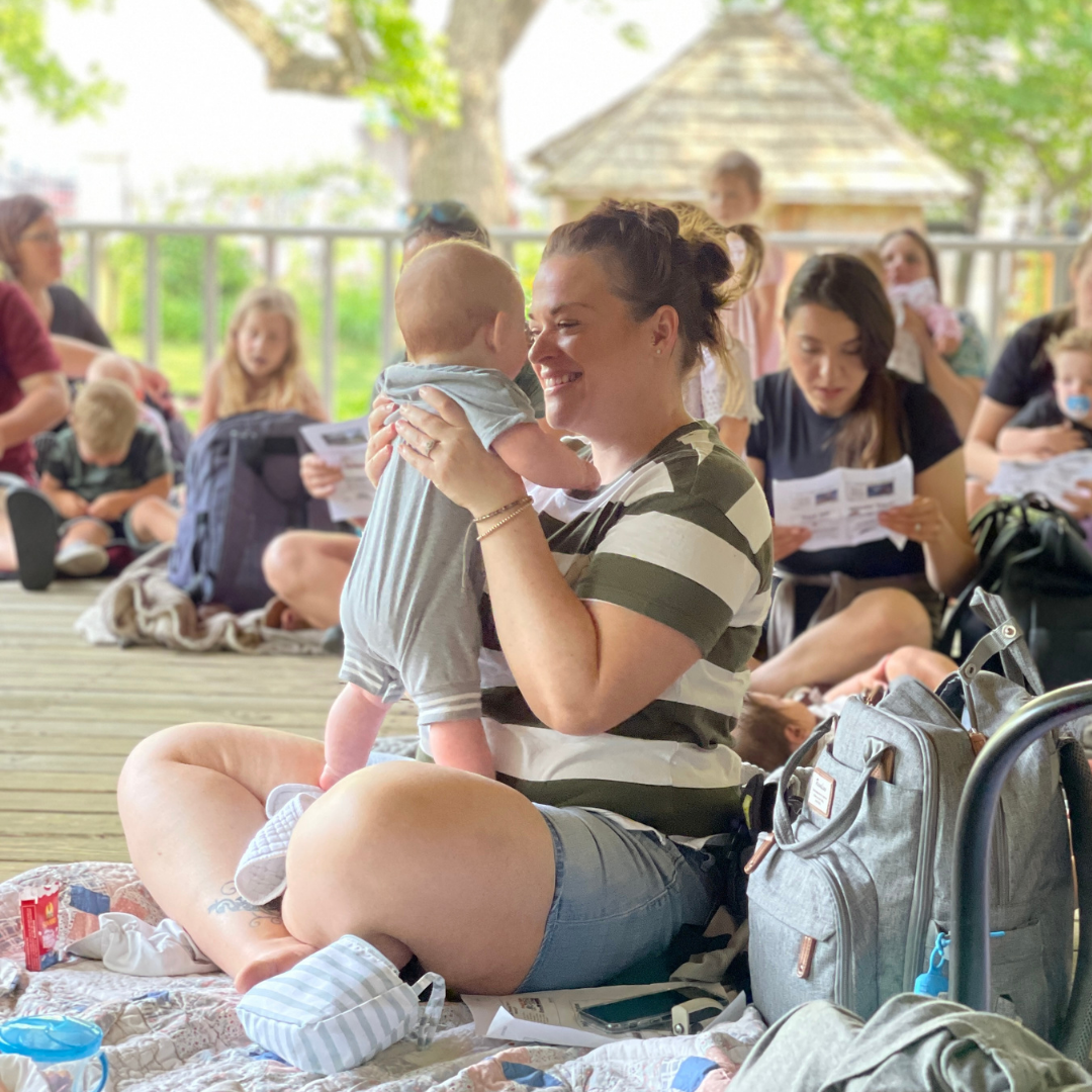A mother smiles while holding her baby during Story Time at Peel Museum on the porch in the late springtime