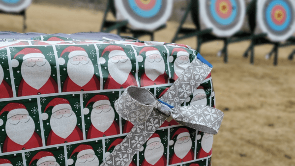 A photo of a box target at The Quiver wrapped in santa claus gift wrapping paper and a silver bow with slow flakes tied around it. Promotes a Christmas themed archery class.
