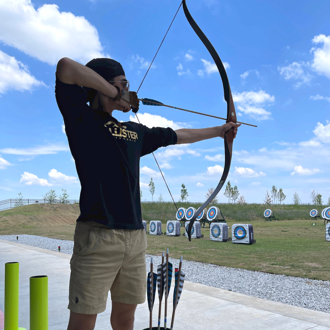 Person shooting a recurve bow with bright blue skies in the background and archery targets behind them in Bentonville, Arkansas