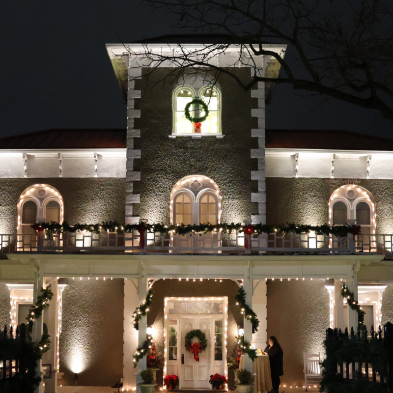 Holiday lights on outside of Victorian Era Historical House and Museum - Peel Museum located in Bentonville Arkansas with greenery around columns and wreaths on door