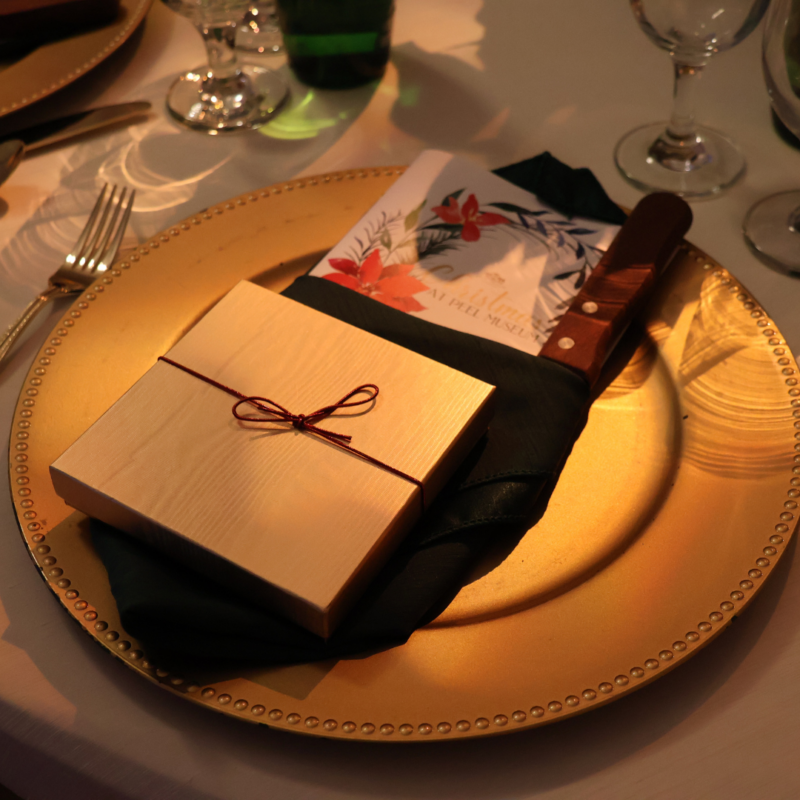 Gold Charger with gift present box on top placed on table setting with Christmas at Peel 2023 Program tucked in napkin