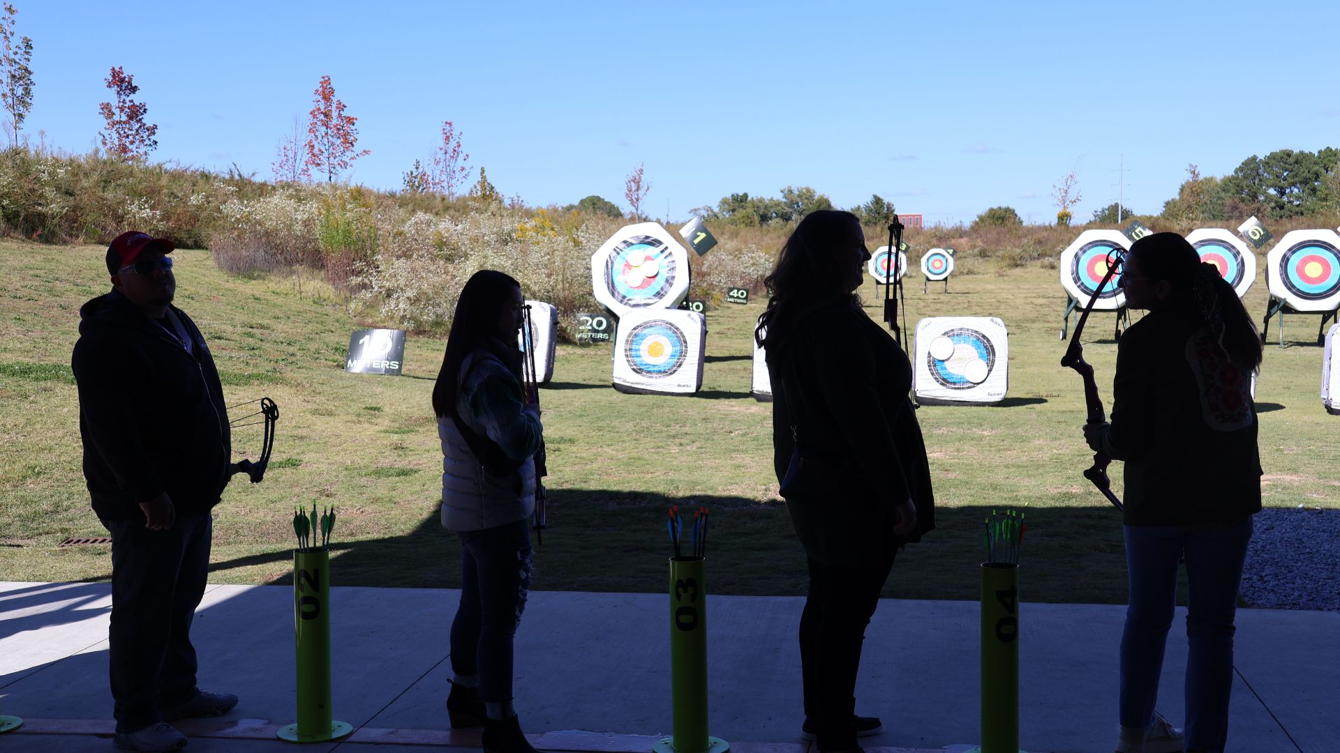 4 silouettes of archers standing in front of outdoor archery range with blue skies at Osage park